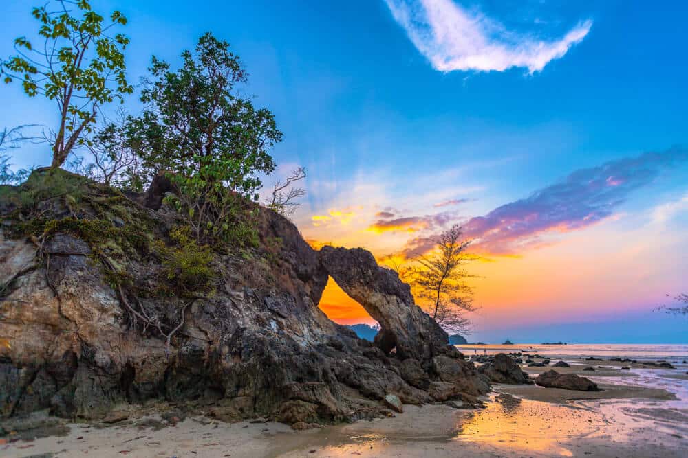 ซุ้มหินธรรมชาติบนชายหาดในช่วงพระอาทิตย์ตกพร้อมท้องฟ้าที่สดใส ที่ท่องเที่ยวระนองแห่งนี้น่าทึ่งมาก  ท่องเที่ยวระนอง ต้นไม้ประดับประดาโขดหิน และเมื่อน้ำลงเผยให้เห็นบริเวณที่มีทรายและเปียกรอบๆ ซุ้มโค้ง