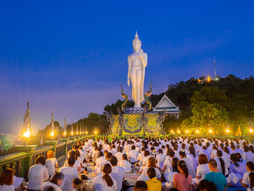 คนจำนวนมากสวมชุดสีขาวนั่งหันหน้าเข้าหาพระพุทธรูปสูงในพิธีตอนเย็นที่วัด โดยมีเนินเขาและต้นไม้เป็นฉากหลัง ซึ่งถือเป็นวันสำคัญ วันมาฆบูชา