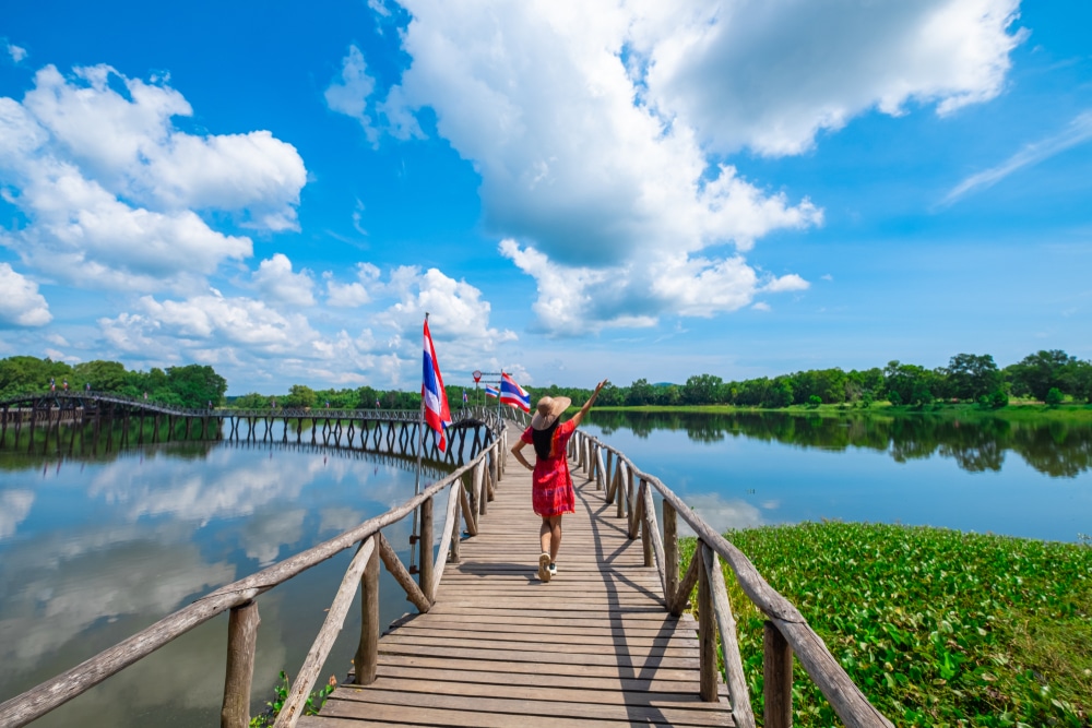 คนสวมชุดสีแดงและหมวกเดินไปตามสะพานไม้เหนือทะเลสาบสะท้อนแสงพร้อมธงและต้นไม้เขียวขจีล้อมรอบบริเวณใต้ท้องฟ้าสีครามสดใสและมีเมฆกระจาย ณ จุดชมวิวเขามัทรี