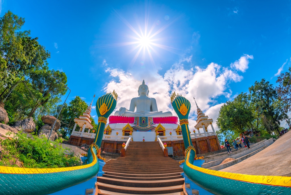 มุมมองแบบ Fisheye ของพระพุทธรูปสีขาวขนาดใหญ่บนแท่นที่มีรูปปั้นพญานาคสีเขียว 2 องค์ขนาบข้างบันได ตัดกับท้องฟ้าที่สดใส ผู้เยี่ยมชมสามารถเห็นเป็นพื้นหลังในการสำรวจสถานที่ท่องเที่ยวขอนแก่นของ ขอนแก่นท่องเที่ยว