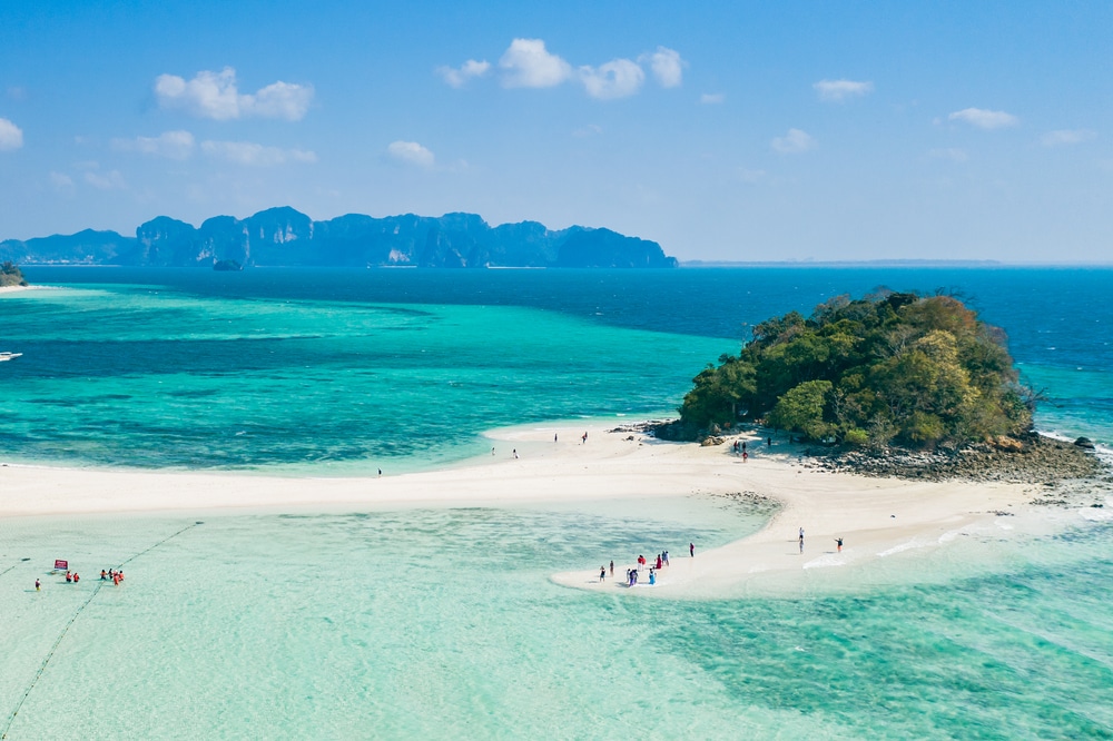 ภาพมุมสูงของเกาะเขตร้อนที่มีหาดทรายสีขาว น้ำทะเลสีฟ้าใส ทะเลใต้สวยๆ และผู้คนกำลังอาบแดด กลุ่มคนเล็กๆ กระจายอยู่บนชายหาดและในน้ำตื้น มองเห็นเนินเขาเขียวขจีอยู่ไกลๆ ทัศนียภาพที่งดงามราวกับอัญมณีแห่งเที่ยวทะเลภาคใต้ ซึ่งดึงดูดนักท่องเที่ยวจำนวนมากตลอดทั้งปี