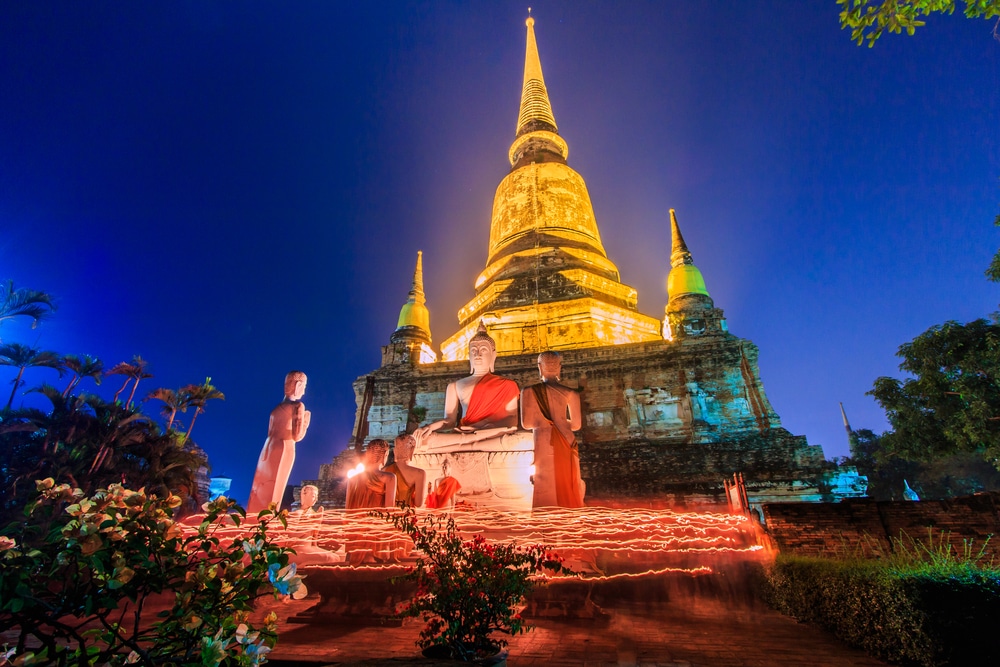 วัดพุทธที่มีแสงสว่างในตอนกลางคืน โดยมีเจดีย์เป็นพื้นหลัง รูปปั้นพระพุทธเจ้าประทับนั่งอยู่ด้านหน้า วันมาฆบูชา  และมีแสงเทียนเป็นวงกลมล้อมรอบรูปปั้นในช่วงวันมาฆบูชา