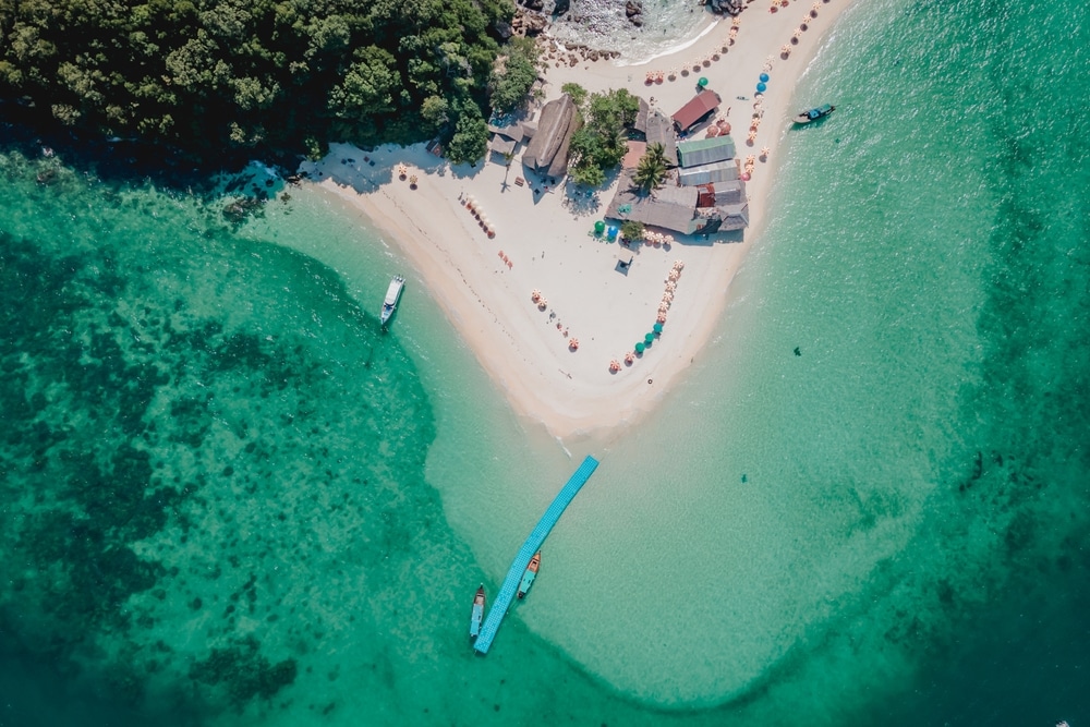 มุมมองทางอากาศของเกาะเขตร้อนที่มีน้ำทะเลสีฟ้าครามใสทะเลใต้สวยๆ  หาดทราย พืชพรรณที่เขียวชอุ่ม อาคารที่มีเสน่ห์ไม่กี่แห่ง เรือ และร่มกันแดด - งดงามที่เที่ยวทะเลใต้