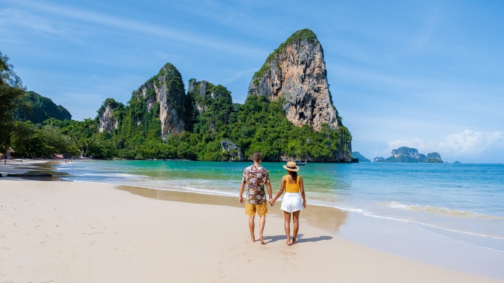 คู่รักเดินจับมือกันไปตามหาดทราย น้ำทะเลสีฟ้าใส และหน้าผาหินปูนสูง ทะเลใต้สวยๆ ตระหง่านภายใต้ท้องฟ้าสีฟ้าสดใส สัมผัสถึงความสวยงามของทะเลใต้อันงดงาม