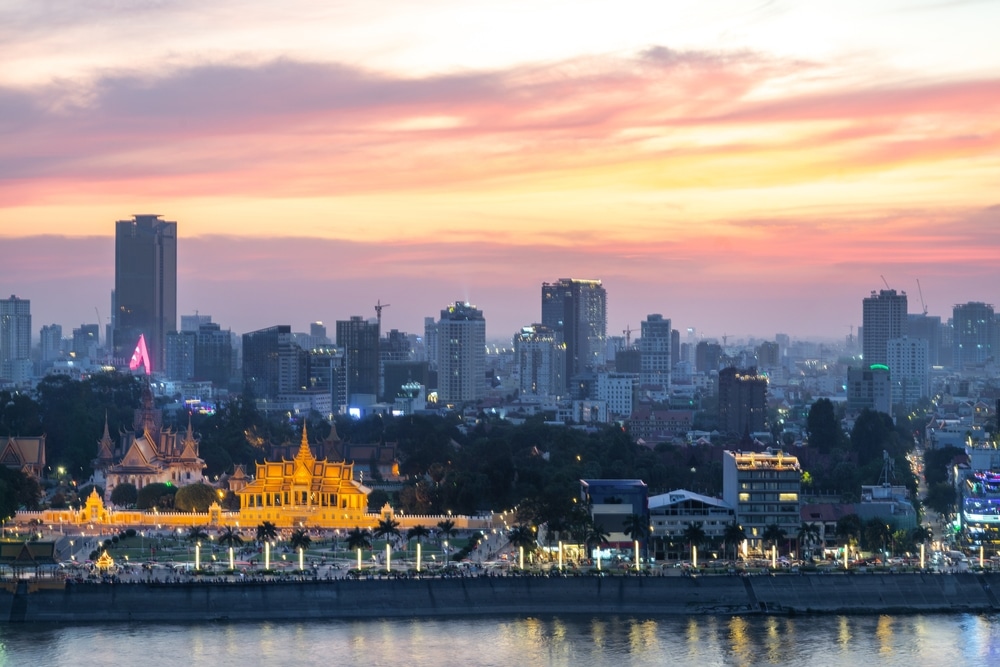 เส้นขอบฟ้าของเมืองยามพลบค่ำพร้อมอาคารที่ส่องสว่าง รวมถึงวัดสีทองที่อยู่เบื้องหน้า โดยมีท้องฟ้ายามพระอาทิตย์ตกหลากสีสัน มีแม่น้ำไหลผ่านด้านล่างของภาพ ทำให้มองเห็นที่เที่ยวพนมเปญได้อย่างน่าหลงใหล