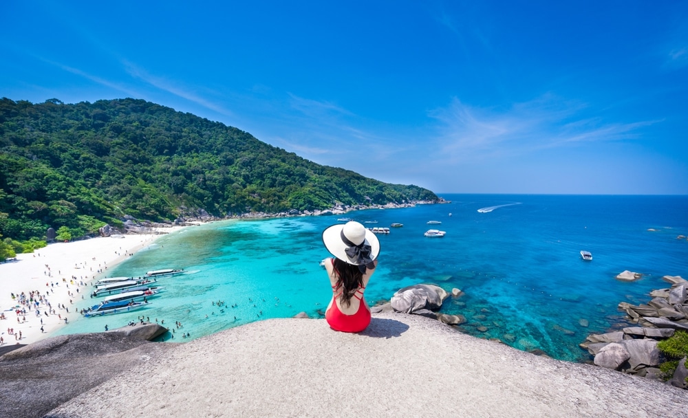 คนสวมชุดสีแดงและหมวกปีกกว้างนั่งอยู่บนก้อนหิน มองเห็นชายหาดที่มีน้ำทะเลสีฟ้าใส เรือ และเนินเขาสีเขียวชอุ่มภายใต้ท้องฟ้าสีฟ้าใส เป็นฉากอันสวยงามที่เหมาะกับใครก็ตามที่ใฝ่ฝันถึง เที่ยวทะเลภาคใต้ .