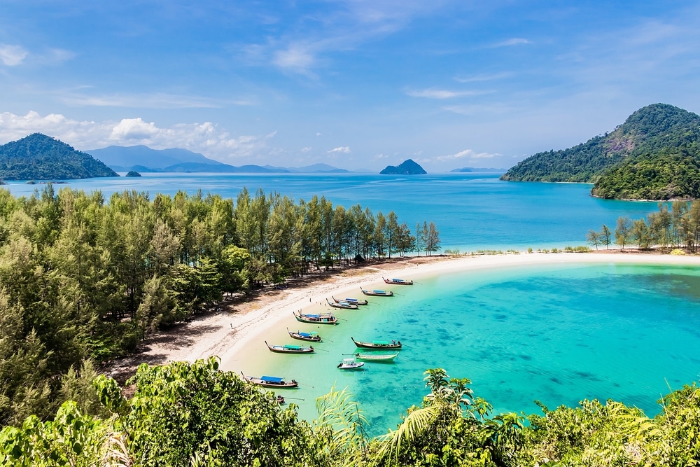 ชายหาดเล็กๆ ท่องเที่ยวระนอง ที่มีเรือเทียบท่าในน้ำสีฟ้าคราม ล้อมรอบด้วยต้นไม้เขียวชอุ่มและเกาะต่างๆ ที่เป็นเนินเขาเป็นพื้นหลังภายใต้ท้องฟ้าสีคราม ถือเป็นจุดหมายปลายทางที่สมบูรณ์แบบสำหรับนักท่องเที่ยวที่ท่องเที่ยวระนอง