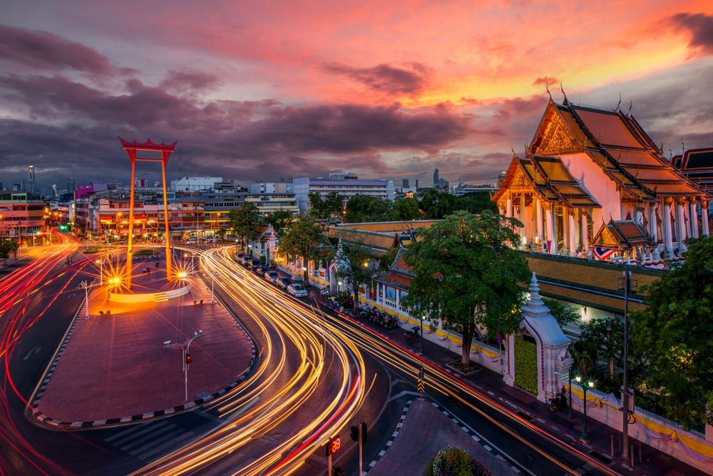 สี่แยกที่พลุกพล่านไปด้วยแสงไฟจากยานพาหนะในยามพลบค่ำ วันมาฆบูชา  แสดงให้เห็นวิหารขนาดใหญ่ที่ประดับประดาอย่างวิจิตรงดงามและประตูพิธีกรรมที่สูงตระหง่านภายใต้ท้องฟ้าที่มีสีสันสดใส สะท้อนถึงจิตวิญญาณของวันมาฆบูชาได้อย่างสวยงาม