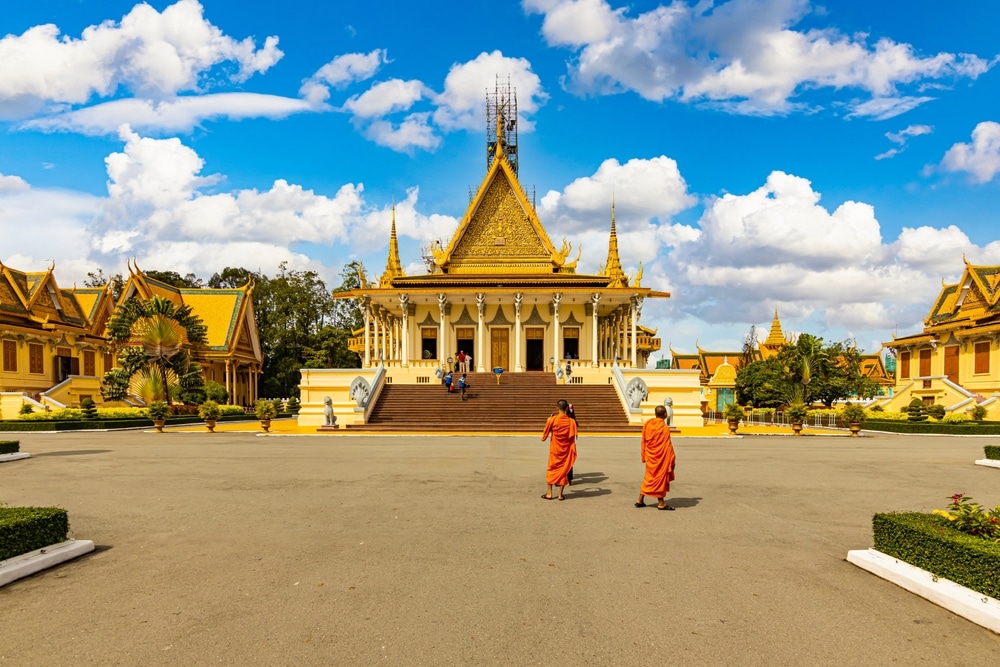 พระภิกษุ 2 รูปในชุดสีส้มเดินไปที่วัด ที่เที่ยวกัมพูชา ขนาดใหญ่ที่ตกแต่งอย่างวิจิตร มียอดแหลมสีทองตัดกับท้องฟ้าสีครามสดใสและมีเมฆ ใกล้บันไดทางเข้ามองเห็นผู้เยี่ยมชมหลายคนได้ เพื่อสำรวจตัวอย่างอันน่าทึ่งของป้ายที่เที่ยว