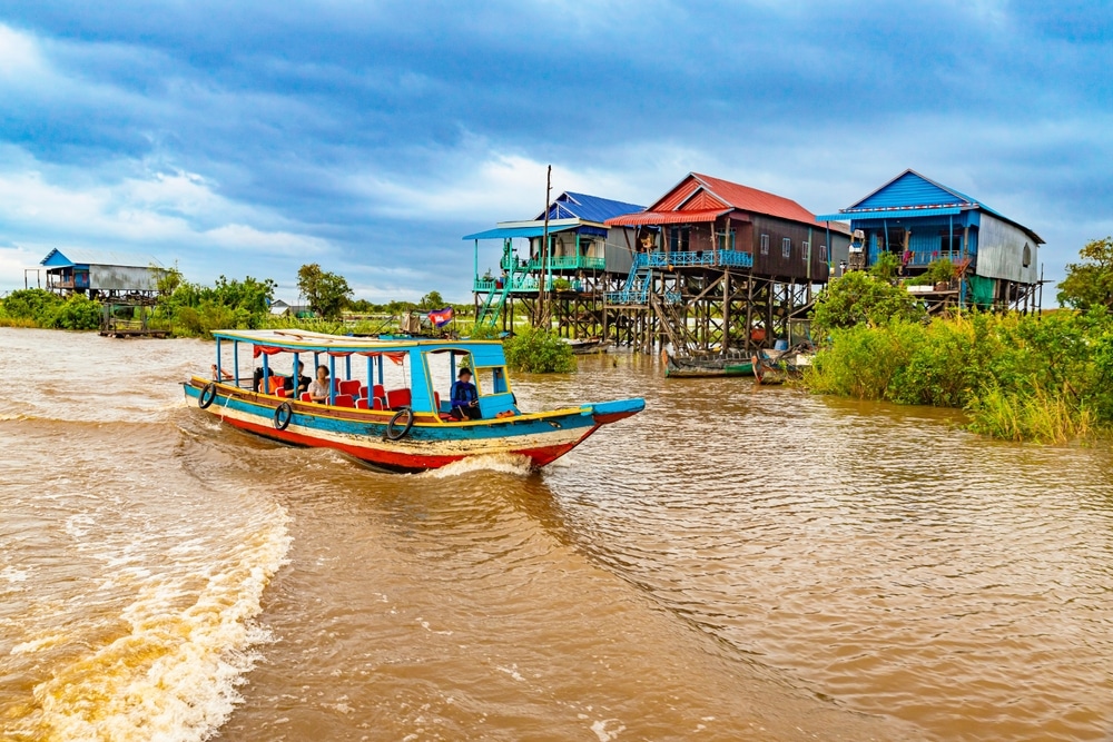 เรือหลากสีสันล่องไปตามแม่น้ำโดยมีบ้านยกสูงเป็นพื้นหลังภายใต้ท้องฟ้าที่มี ที่เที่ยวกัมพูชา เมฆมาก เผยให้เห็นถึงเสน่ห์ของกระดาษแข็งที่เที่ยว