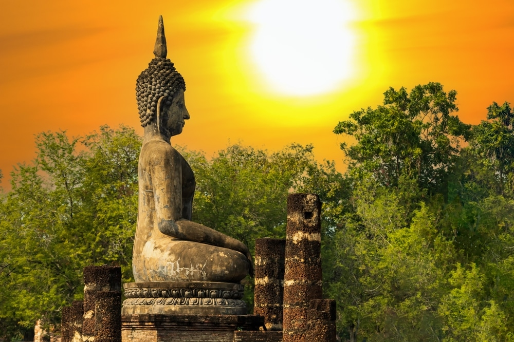 พระพุทธรูปหินประทับนั่งในท่าสมาธิท่ามกลางซากปรักหักพังโบราณสถาน โดยมีพระอาทิตย์ตกสีส้มสดใสและต้นไม้สีเขียวชอุ่มอยู่เบื้องหลัง สื่อถึงจิตวิญญาณแห่งการ สวดมนต์ข้ามปี ขณะที่ธรรมชาติและประวัติศาสตร์บรรจบกันอย่างกลมกลืนและเงียบสงบ