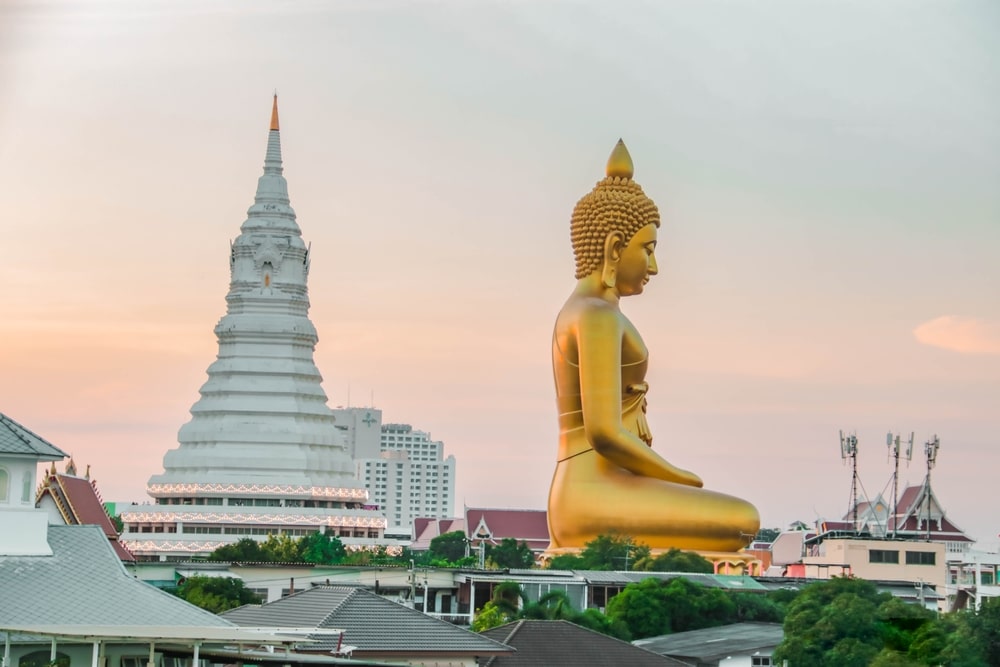 พระพุทธรูปสีทององค์ใหญ่ประดิษฐานอยู่หน้าเจดีย์สีขาว ท่ามกลางอาคารในเมืองและต้นไม้สีเขียว ภายใต้ท้องฟ้าสีชมพูอ่อนและสีส้ม สร้างฉากหลังอันเงียบสงบให้กับงานเฉลิมฉลอง วันมาฆบูชา