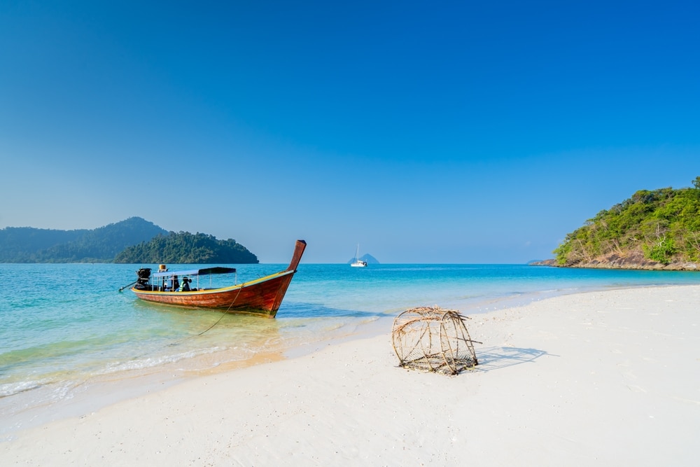 เรือหางยาวจอดทอดสมออยู่ใกล้หาดทรายขาว น้ำทะเลสีฟ้าใส ท่องเที่ยวระนอง มีเกาะสีเขียวเป็นฉากหลัง และมีกับดักปลาวางอยู่บนฝั่ง ฉากอันเงียบสงบนี้เป็นตัวอย่างที่ดีที่สุดของระนอง