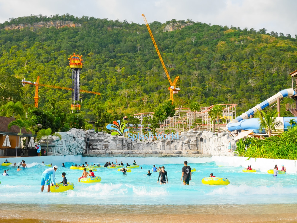 ผู้คนกำลังเพลิดเพลินกับสระน้ำมีคลื่นที่สวนน้ำ Splash World โดยมีเนินเขาเขียวขจีและมีเครนก่อสร้างหลายตัวอยู่เบื้องหลัง เป็นจุดที่เหมาะสำหรับพาลูกเที่ยวธรรมชาติใกล้กรุงเทพ มีทั้งความสนุกสนานและทิวทัศน์ที่สวยงาม ขับรถเพียงไม่นานจากตัวเมือง Places to visit zoos near Bangkok