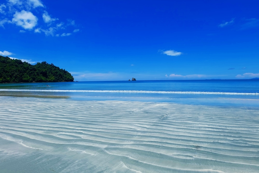 ชายหาดอันบริสุทธิ์ที่มีหาดทรายที่ใสระลอกคลื่น คลื่นที่อ่อนโยน และ ท่องเที่ยวระนอง ท้องฟ้าสีครามกำลังรอคอยที่ท่องเที่ยวระนองที่สวยงามแห่งนี้ มองเห็นเนินเขาสีเขียวทางด้านซ้าย และมีกลุ่มหินเล็กๆ อยู่ในระยะไกล