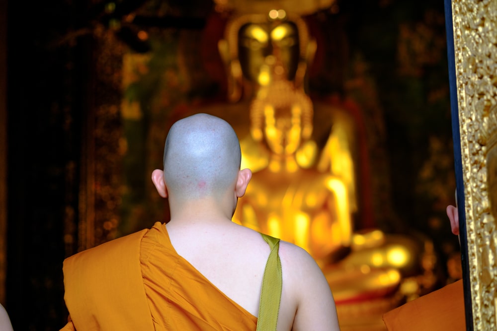 พระภิกษุโกนหัวสวมจีวรสีส้มยืนหันหน้าเข้าหาพระพุทธรูปสีทององค์ใหญ่ในร่มอย่างเงียบๆ ขณะสังเกตความสงบของ วันมาฆบูชา