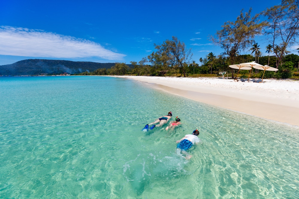 คนสามคนดำน้ำดูปะการังในทะเลสีฟ้าครามใกล้ ที่เที่ยวกัมพูชา หาดทรายขาวที่มีต้นไม้และร่มเป็นพื้นหลังภายใต้ท้องฟ้าสีฟ้าใส เป็นวันที่เหมาะสำหรับการเพลิดเพลินไปกับสถานที่ท่องเที่ยวที่สวยงามแห่งหนึ่ง