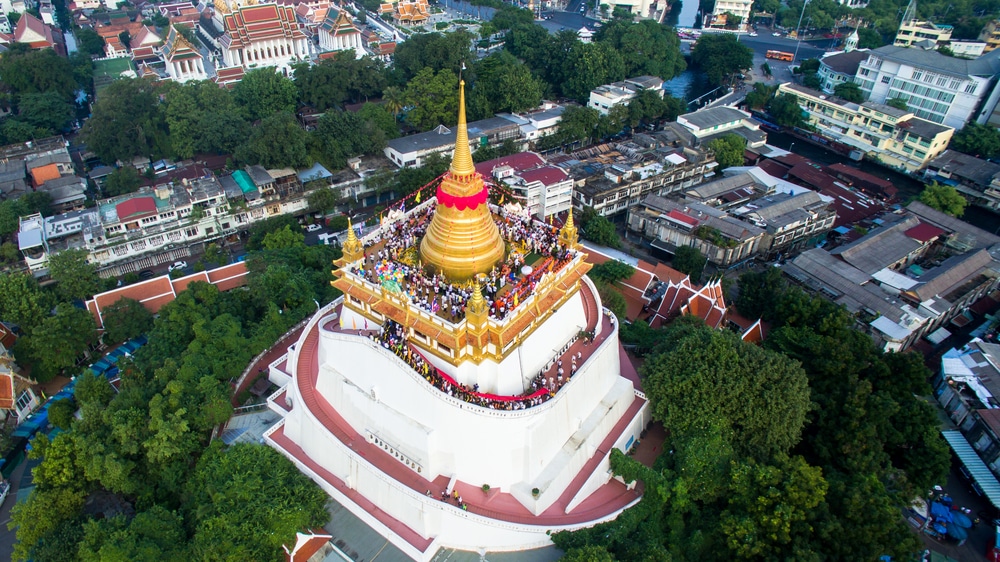 ภาพมุมสูงของวัดภูเขาทองในกรุงเทพมหานคร ประเทศไทย มีผู้คนจำนวนมากมารวมตัวกันรอบ ๆ เจดีย์ทองเพื่อเฉลิมฉลอง วันมาฆบูชา มองเห็นทิวทัศน์เมืองและต้นไม้โดยรอบเป็นฉากหลัง