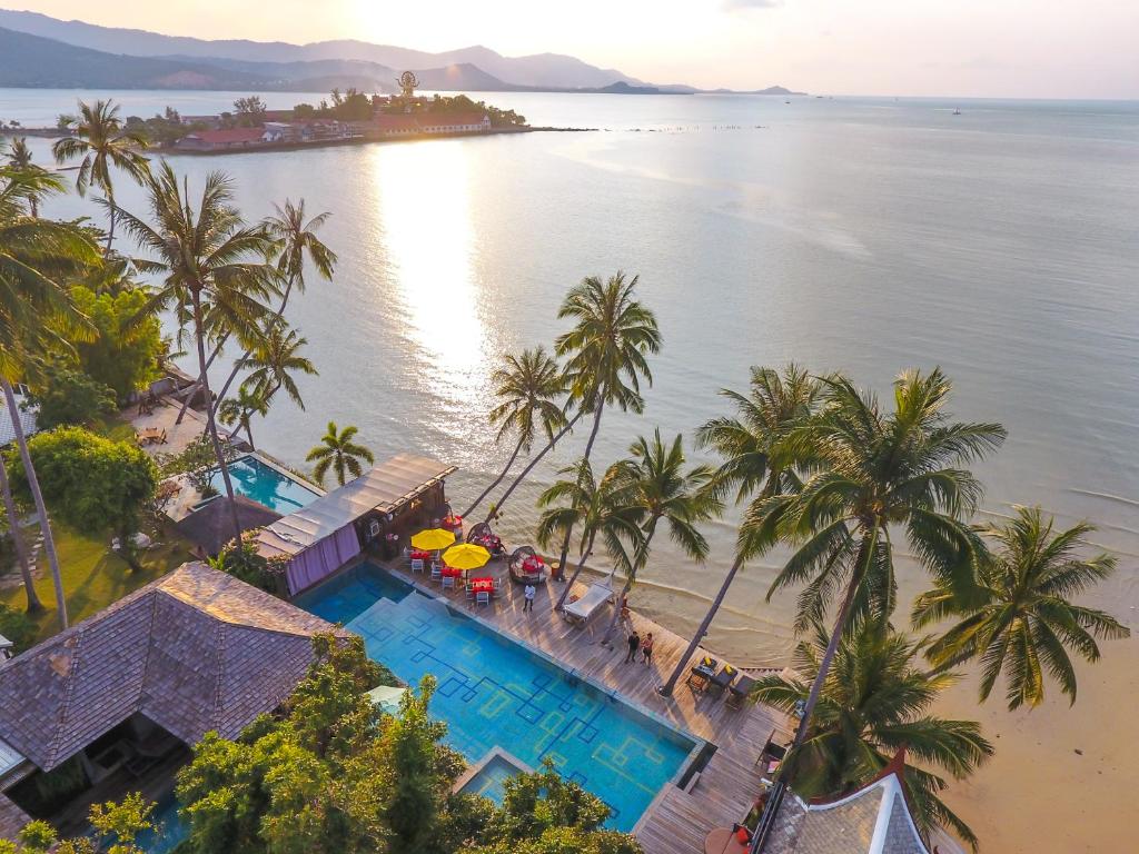 มุมมองทางอากาศของรีสอร์ทริม ที่พักวิวทะเล ชายหาดอันเงียบสงบที่มีต้นปาล์ม สระว่ายน้ำขนาดใหญ่ และทะเลอันเงียบสงบภายใต้ท้องฟ้าที่แจ่มใส โดยมีเนินเขาที่มองเห็นได้ในระยะไกล—สถานที่พักผ่อนที่สมบูรณ์แบบ