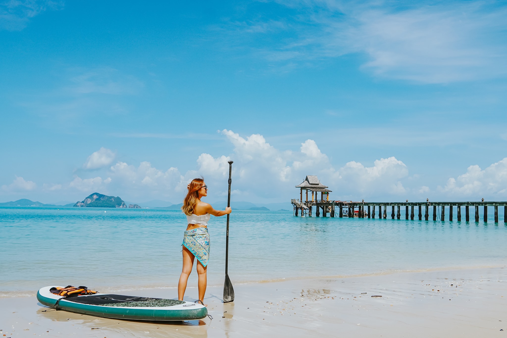 ผู้หญิงคนหนึ่งสวมชุดว่ายน้ำ ที่พักวิวทะเล และผ้าถุงยืนอยู่บนชายหาดทราย ถือไม้พายไว้ข้างกระดานพาย เธอมองไปยังท่าเทียบเรือและทะเลอันสงบ และเพลิดเพลินกับการพักผ่อนใต้ท้องฟ้าใสที่มีเมฆกระจัดกระจาย