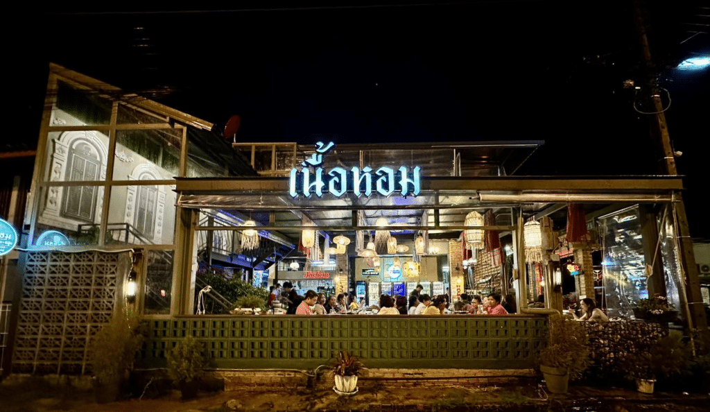 บรรยากาศยามค่ำคืนของร้านอาหารที่คึกคักพร้อมป้ายไฟที่เขียนด้วยอักษรต่างประเทศ ลูกค้ากำลังรับประทาน ที่ท่องเที่ยวอุบลราชธานี อาหารใต้แสงอุ่นๆ และร้านอาหารแห่งนี้ได้รับการออกแบบแบบเปิดโล่งในสไตล์ชนบท