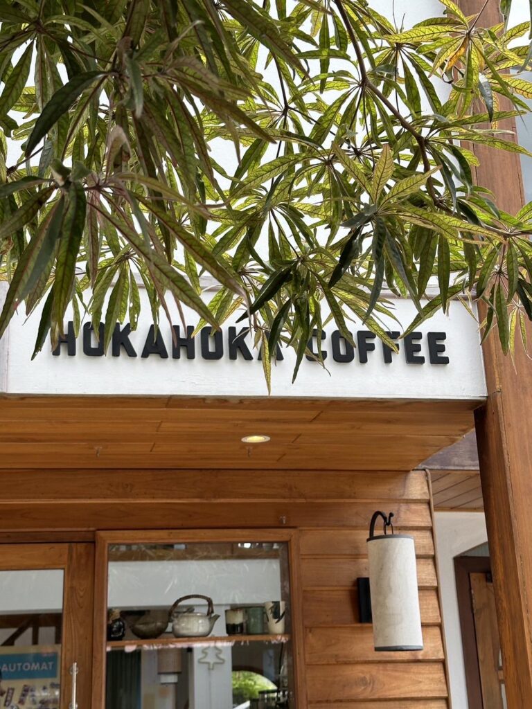 ป้ายที่ติดอยู่ภายนอกอาคารไม้เขียนว่า “Huka Huka Coffee” โดยมีใบไม้สีเขียวบดบังทัศนียภาพบางส่วน ท่องเที่ยวอุบลราชธานี