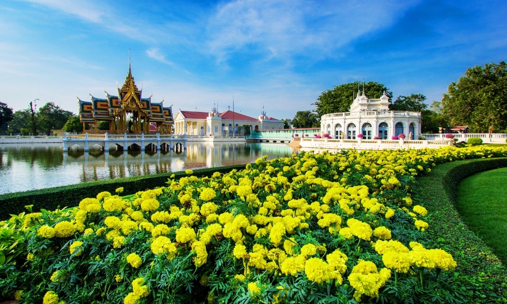 สวนภูมิทัศน์ที่มีดอกไม้สีเหลืองสดใสอยู่เบื้องหน้า สระน้ำใส และ ท่องเที่ยวอยุธยา สถาปัตยกรรมไทยที่หรูหราภายใต้ท้องฟ้าสีครามในพื้นหลัง รวบรวมแก่นแท้ของการท่องเที่ยวจังหวัดอยุธยา