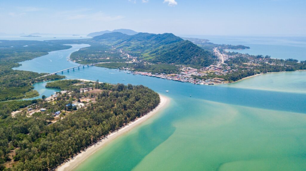 มุมมองทางอากาศของพื้นที่ชายฝั่งทะเลที่มีชายหาด สถานที่ท่องเที่ยวจังหวัดกระบี่ แม่น้ำ สะพานเชื่อมแผ่นดิน และเนินเขาสีเขียวเป็นฉากหลัง