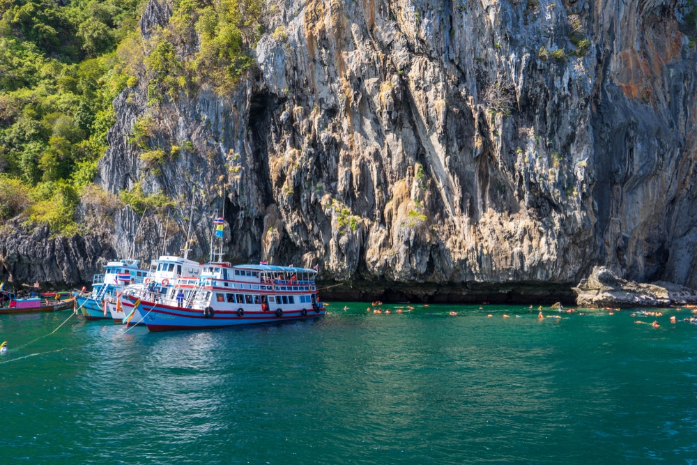 เรือลำใหญ่จอดทอดสมออยู่ใกล้หน้าผาหินที่มีต้นไม้เขียวขจี เป็นสถานที่สำหรับการดำน้ำที่น่าตื่นเต้น  ทริปดำน้ำ ผู้คนจำนวนมากกำลังว่ายน้ำและดำน้ำตื้นในน้ำสีฟ้าอมเขียวใส เพลิดเพลินไปกับโลกใต้ทะเลที่สดใส