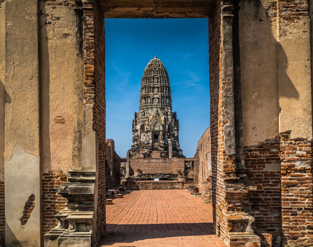 มองผ่านซุ้มประตูหินของวัดประวัติศาสตร์ในอยุธยาที่มียอดแหลมแกะสลักอย่าง ท่องเที่ยวอยุธยา วิจิตรงดงามตัดกับท้องฟ้าสีฟ้าสดใส