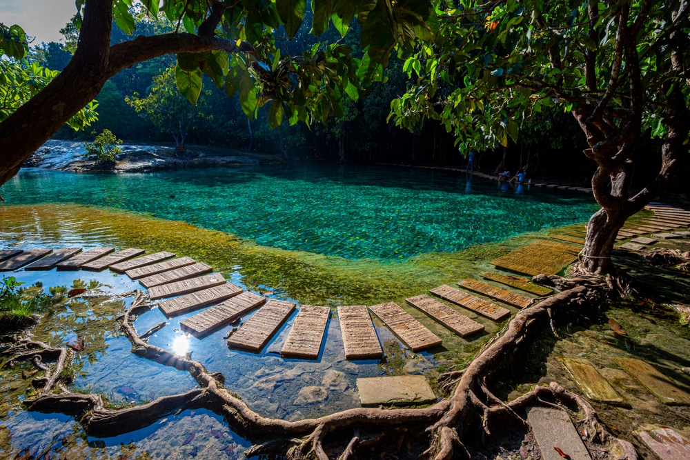 น้ำทะเลสีฟ้าใสที่มีขั้นบันไดหินนำไปสู่สระน้ำธรรมชาติ 10 สถานที่ท่องเที่ยวกระบี่ ล้อมรอบด้วยต้นไม้และใบไม้ มีแสงแดดสะท้อนบนผิวน้ำ