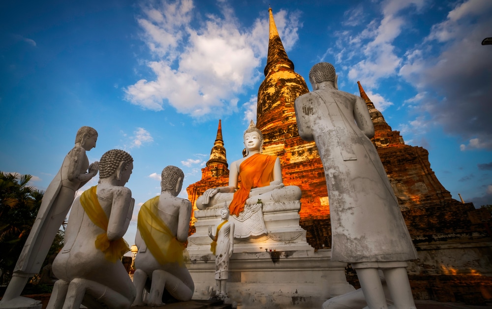 รูปปั้นพระพุทธเจ้าและผู้ติดตามในวัดกลางแจ้งที่มีเจดีย์ขนาดใหญ่เป็นพื้นหลังภายใต้ท้องฟ้าสีครามและมีเมฆ ทำให้เกิดเป็นฉากที่งดงามสมบูรณ์แบบสำหรับสถาปัตยกรรมอยุธยา