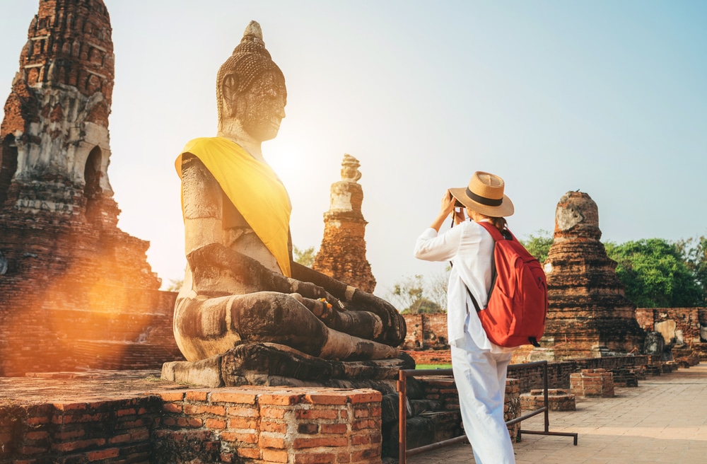 ผู้สวมหมวกและกระเป๋าเป้สีแดงถ่ายภาพพระพุทธรูปปางสมาธิขนาดใหญ่ที่ห่อหุ้มผ้าสีเหลืองในวัดโบราณภายใต้ท้องฟ้าที่แจ่มใส ท่องเที่ยวอยุธยา รวบรวมแก่นแท้ของสถาปัตยกรรมอยุธยา