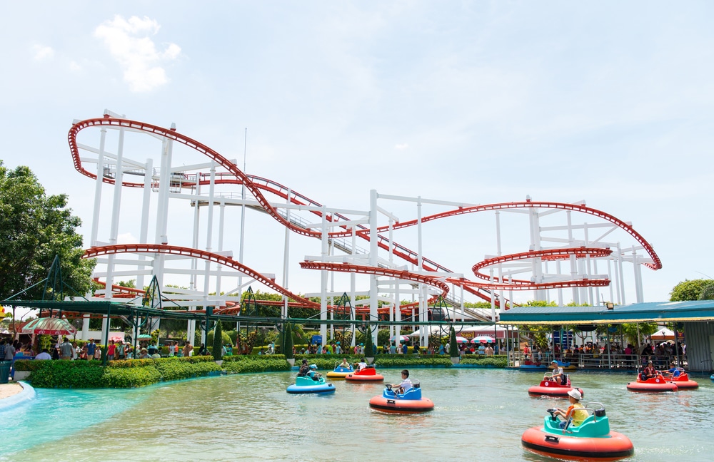 ผู้คนนั่งเรือบัมพ์ในสระน้ำหน้ารถไฟเหาะสีแดงขาวในวันที่อากาศแจ่มใส เหมาะสำหรับผู้ที่กำลังมองหาที่เที่ยวหน้าฝนโรแมนติก ที่เที่ยวครอบครัว