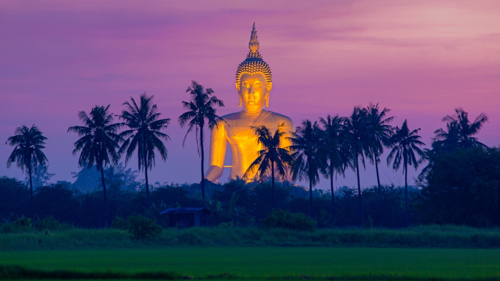 พระพุทธรูปเรืองแสงขนาดใหญ่ตั้งอยู่ท่ามกลางต้นปาล์มในเวลาพลบค่ำ ใต้ท้องฟ้าสีม่วงและสีชมพู ซึ่งเป็นภาพที่น่าหลงใหลซึ่งเหมาะสำหรับผู้ที่สำรวจอัญมณีที่ไม่ค่อยมีคนรู้จักของ ท่องเที่ยวเมืองรอง ชวนเพื่อน ชเมืองรอง