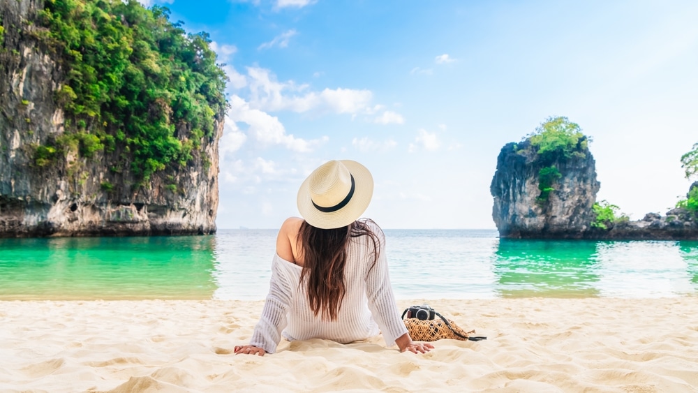 บุคคลสวมชุดสีขาวและหมวกนั่งอยู่บนชายหาดทรายหันหน้า 10 สถานที่ท่องเที่ยวกระบี่ ไปทางน้ำทะเลสีฟ้าใส มีเกาะหินและพืชพรรณสีเขียวเป็นพื้นหลังภายใต้ท้องฟ้าสีฟ้า