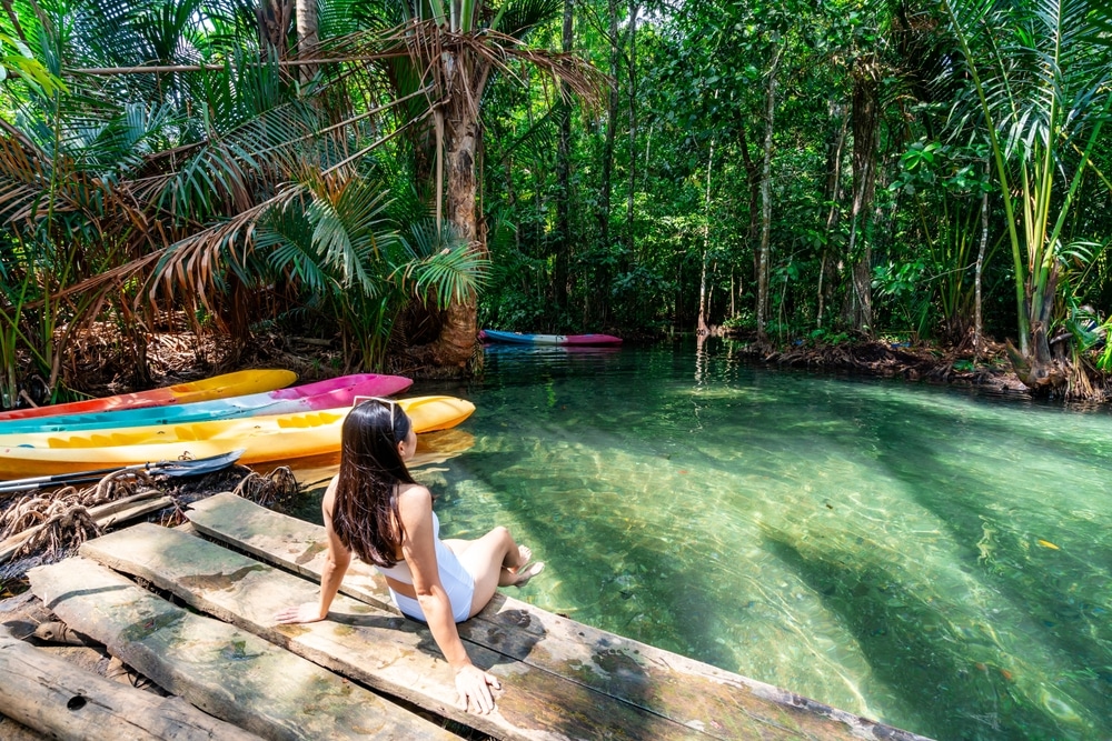 ผู้หญิงคนหนึ่งนั่งอยู่บนท่าเรือไม้ข้างลำธารในป่าที่ใสสะอาด 10 สถานที่ท่องเที่ยวกระบี่ และมีเรือคายัคสีสันสดใสหลายลำอยู่ฝั่งตรงข้าม