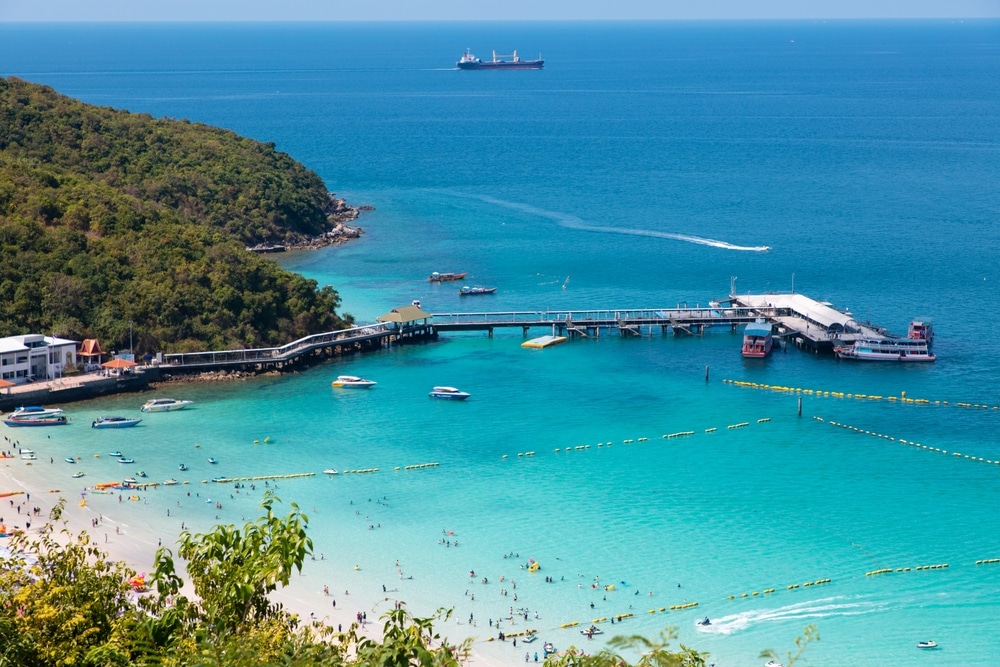 วิวทางอากาศของชายหาดที่มีน้ำทะเลสีฟ้าใส ท่าเรือที่มีเรือเทียบท่า และมีเรือขนาดใหญ่อยู่ไกลๆ ภูมิทัศน์อันเขียวชอุ่มล้อมรอบบริเวณ ทำให้เป็นสถานที่ที่เหมาะสำหรับคู่รักที่กำลังมองหาสถานที่พักผ่อน ที่เที่ยวครอบครัว