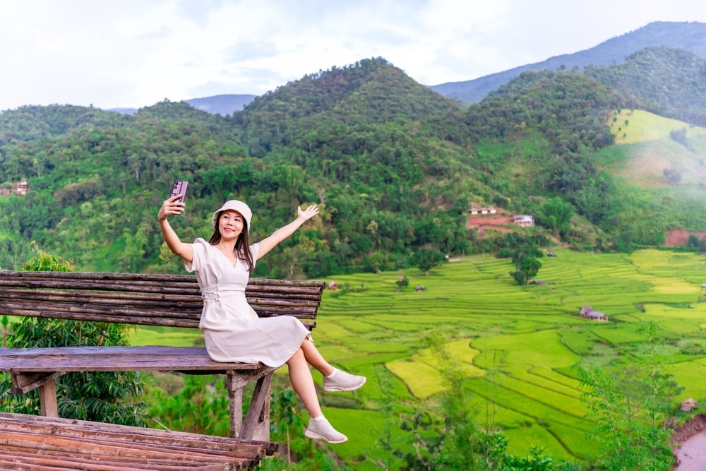 ผู้หญิงสวมชุดสีขาวและหมวกนั่งอยู่บนม้านั่งไม้ ถ่ายเซลฟี่โดยกางแขนออก มองไปยังหุบเขาและภูเขาสีเขียวชอุ่ม ที่ท่องเทียวน่าน