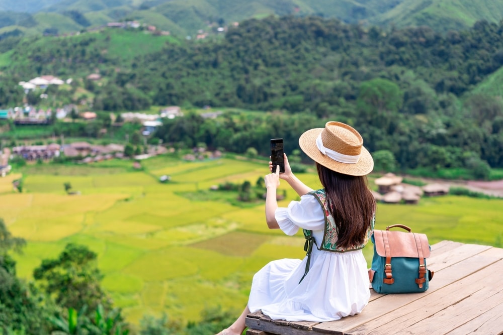 ผู้หญิงสวมชุดสีขาวและหมวกฟางนั่งบนแท่นไม้ถ่ายรูปด้วยโทรศัพท์  ท่องเที่ยวน่าน เธอมองออกไปเห็นหุบเขาสีเขียวและภูเขา มีเป้สะพายหลังวางอยู่ข้างๆ เธอ