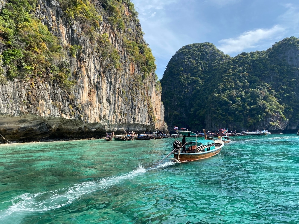 เรือไม้แบบดั้งเดิมแล่นผ่านน้ำสีฟ้าใสใกล้หน้าผาสีเขียวสูงตระหง่าน เหมาะสำหรับการดำน้ำ เรือหลายลำจอดอยู่ใกล้กับแนวชายฝั่งหิน ทริปดำน้ำ เป็นฉากหลังภายใต้ท้องฟ้าสดใส