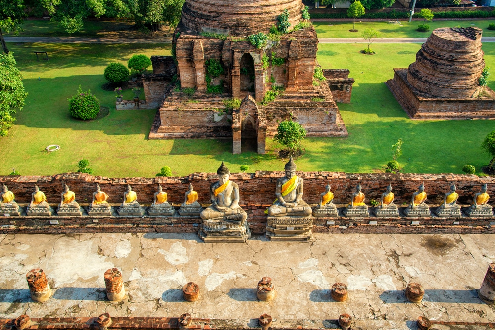 มุมมองทางอากาศของกลุ่มวัดโบราณในอยุธยา ท่องเที่ยวอยุธยา ซึ่งมีพระพุทธรูปหลายองค์คลุมผ้าสีเหลืองและโครงสร้างอิฐโบราณที่ผุกร่อน ล้อมรอบด้วยสนามหญ้าและต้นไม้สีเขียว