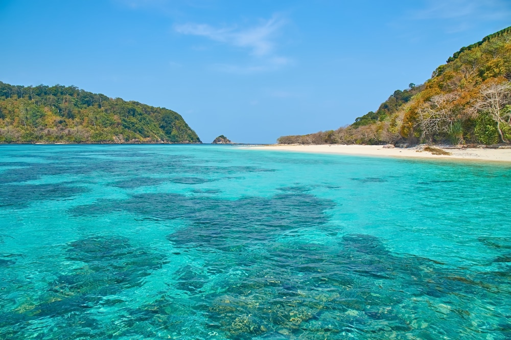 ชายหาดที่เงียบสงบ เหมาะสำหรับการดำดำน้ำ มีน้ำทะเลสีฟ้าใส ทริปดำน้ำ ล้อมรอบด้วยหน้าผาหินสีเขียว และทรายสีขาวภายใต้ท้องฟ้าสีฟ้าใส