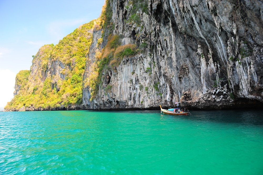 เรือลำเล็กลอยอยู่ใกล้หน้าผาหิน ทริปดำน้ำ ขนาดใหญ่ข้าง ทริปดำน้ำ น้ำทะเลสีฟ้าใสราวกับคริสตัล เป็นจุดที่สมบูรณ์แบบสำหรับการดำน้ำ ต้นไม้เขียวชอุ่มปกคลุมยอดผา ตัดกับท้องฟ้าใสสดใสอย่างสวยงาม