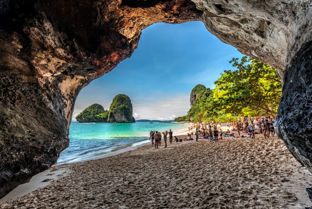 ฉากชายหาดที่มองจากภายในถ้ำ แสดงให้เห็นน้ำทะเลสีฟ้าใส 10 สถานที่ท่องเที่ยวกระบี่ ชายหาดทราย ต้นไม้เขียวขจี และกลุ่มผู้คนที่กำลังเพลิดเพลินกับแสงแดด