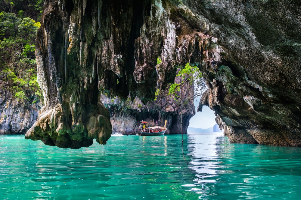 เรือไม้ที่มีหลังคาสีแดงล่องไป 10 สถานที่ท่องเที่ยวกระบี่ ตามน้ำสีฟ้าครามอันเงียบสงบใต้หินปูนขนาดใหญ่และต้นไม้เขียวขจีในสภาพแวดล้อมคล้ายถ้ำ