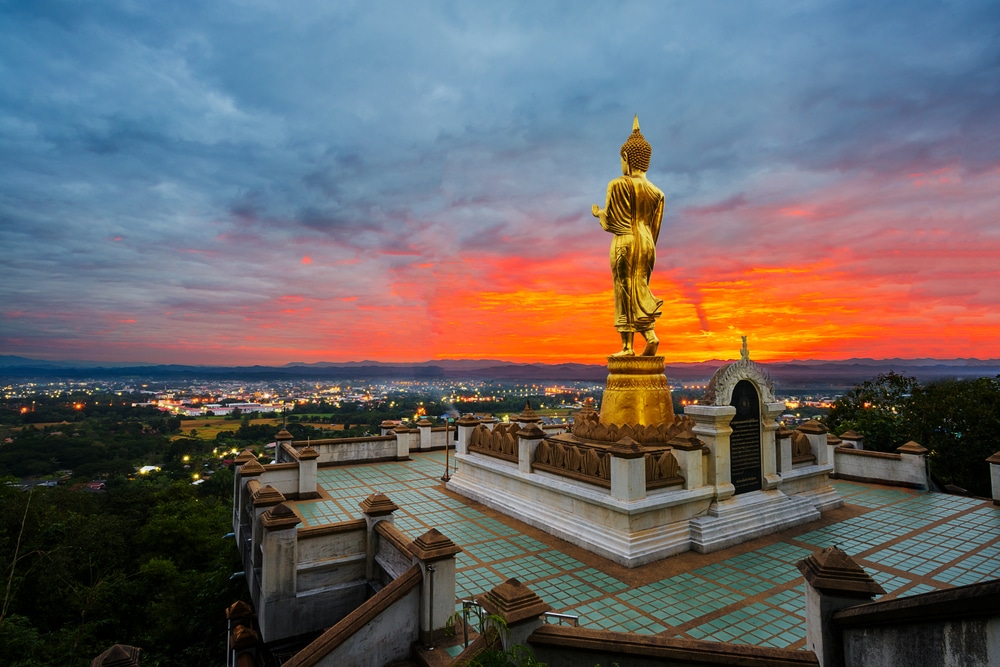 พระพุทธรูปสีทองตั้งตระหง่านท่ามกลางทัศนียภาพเมืองยาม ท่องเที่ยวน่าน พระอาทิตย์ตกดิน โดยมีเมฆสีส้มและสีแดงสดใสบนท้องฟ้า พระพุทธรูปตั้งอยู่บนพื้นที่ยกพื้นพร้อมบันไดและราวบันได