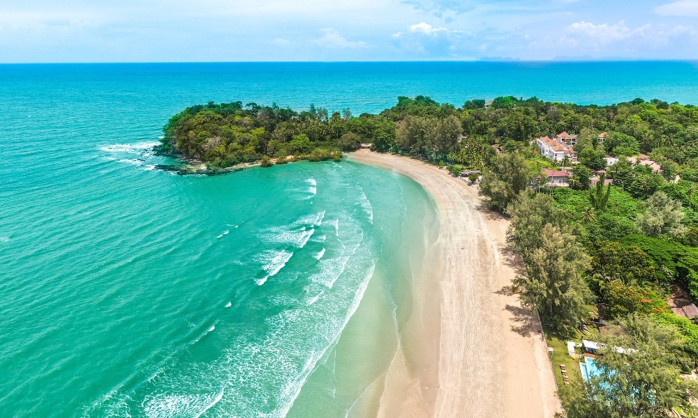 มุมมองทางอากาศของชายหาดที่เงียบสงบ น้ำทะเลสีฟ้าใส 10 สถานที่ท่องเที่ยวกระบี่ คลื่นเบาๆ ต้นไม้สีเขียวขจี และท้องฟ้าสีฟ้าใส สามารถมองเห็นอาคารที่อยู่อาศัยท่ามกลางต้นไม้ใกล้ชายฝั่ง