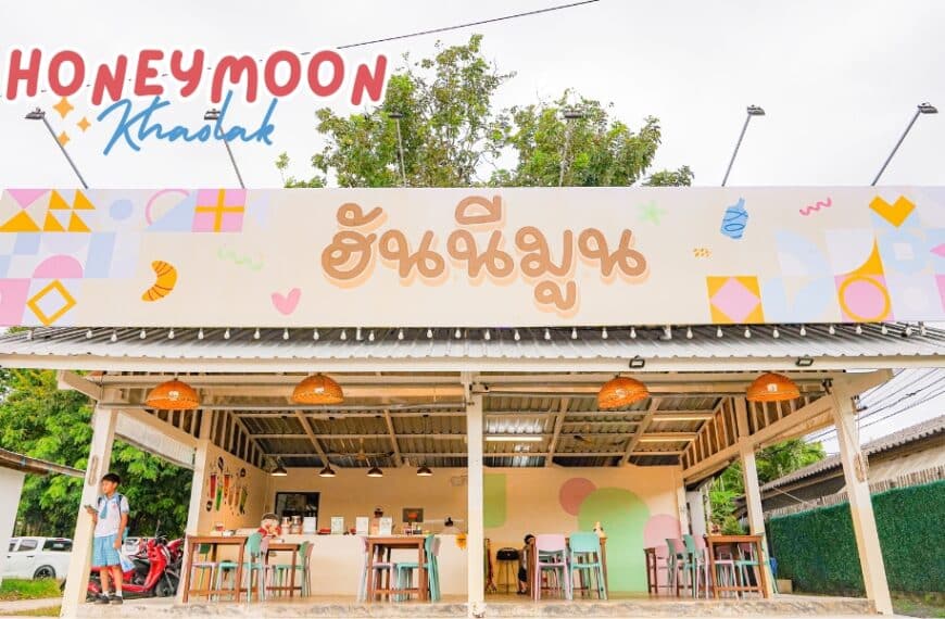 ภายนอกของร้าน Honeymoon cafe ที่มีชีวิตชีวามีป้ายขนาดใหญ่เขียนว่า "Honeymoon Khaolak" มีโต๊ะและเก้าอี้จัดวางไว้ด้านนอก โดยมีคนสองคนยืนรออยู่ใกล้ทางเข้าที่น่าดึงดูด