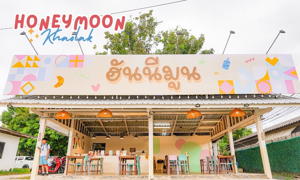 ภายนอกของร้าน Honeymoon cafe ที่มีชีวิตชีวามีป้ายขนาดใหญ่เขียนว่า "Honeymoon Khaolak" มีโต๊ะและเก้าอี้จัดวางไว้ด้านนอก โดยมีคนสองคนยืนรออยู่ใกล้ทางเข้าที่น่าดึงดูด